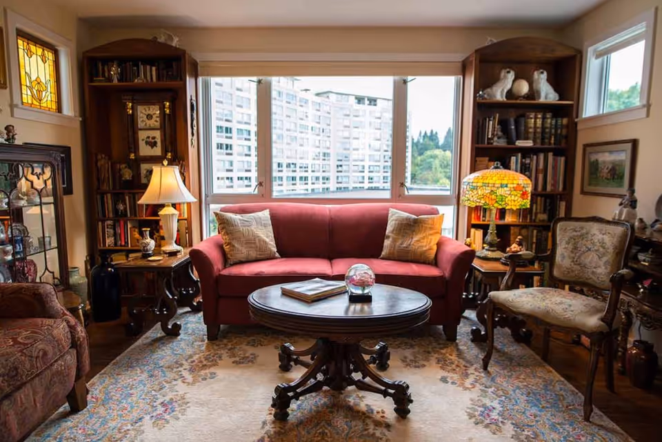 A cozy living room with a red sofa adorned with two patterned pillows, a round wooden coffee table with a glass decorative item and a book, two wooden bookshelves filled with books and decorative items, a stained glass table lamp, an armchair with floral upholstery, and a large window showing a view of a building and greenery outside.