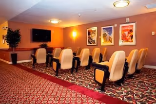 Small theater-style common room with two rows of upholstered recliner chairs facing a wall-mounted TV, framed artwork, and warm lighting.