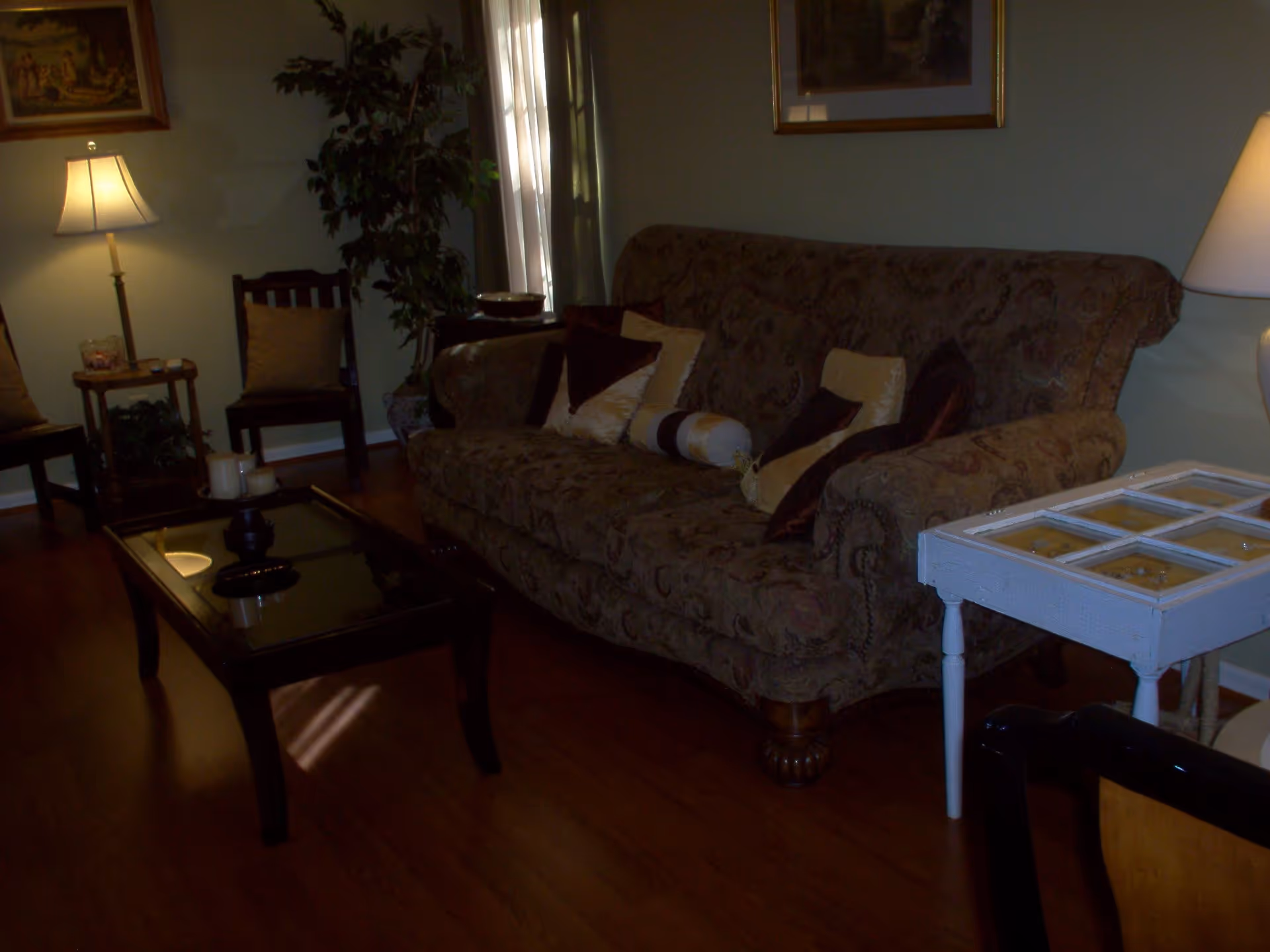A cozy living room with a patterned sofa adorned with multiple decorative pillows, a glass-top coffee table with candles, two wooden chairs, a floor lamp, a potted plant, and framed artwork on the walls.