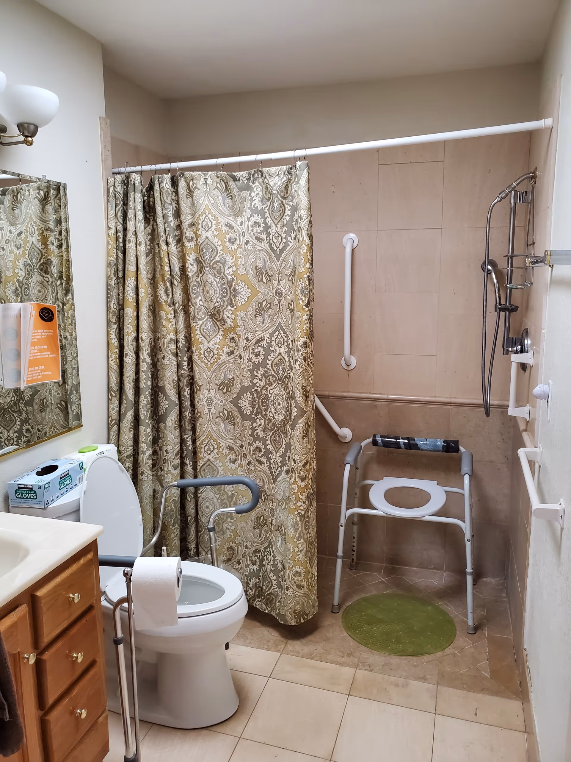 A bathroom with accessibility features including grab bars near the toilet and shower area, a shower chair, a patterned shower curtain, a toilet with arm supports, a sink with wooden cabinets, and a box of gloves on the toilet tank.