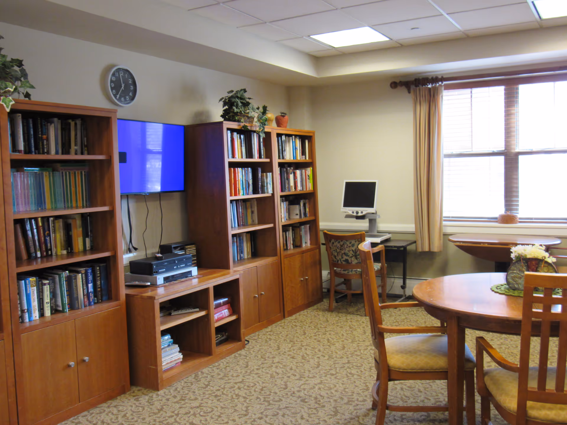 A cozy room with wooden bookshelves filled with books, a wall-mounted TV, a clock above the TV, a computer on a desk with a chair, a round wooden table with chairs, and a window with beige curtains letting in natural light.