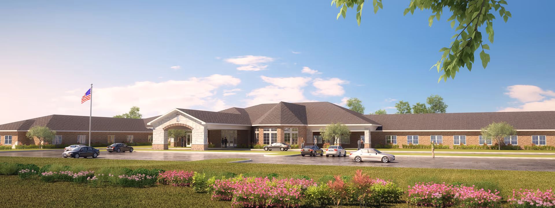 Front exterior view of a single-story senior living facility with a large covered entrance, several cars parked in front, an American flag on a flagpole, and landscaped grounds with green grass and flowering bushes under a partly cloudy sky.