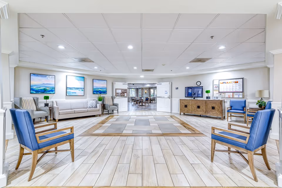 A spacious and well-lit senior living facility common area with blue and beige chairs arranged around the perimeter. There is a beige sofa and two armchairs against the left wall, three landscape paintings above the sofa, and a wooden cabinet with a TV and clock on the right side. Double doors in the center lead to a dining area with tables and chairs.