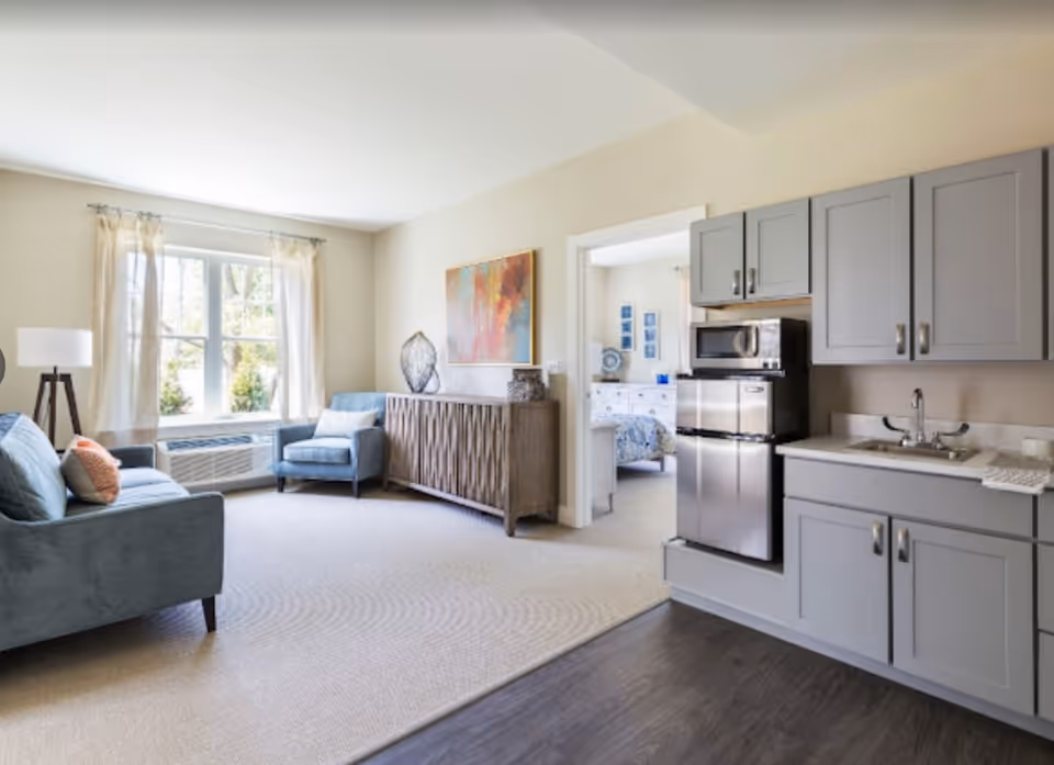 A bright and airy living area with a large window covered by sheer curtains, a blue sofa with pillows, a matching armchair, a wooden sideboard with decorative items, and a colorful abstract painting on the wall. To the right, there is a kitchenette with gray cabinets, a small refrigerator, a microwave, and a sink. Through an open doorway, a bedroom with a bed and dresser is visible.