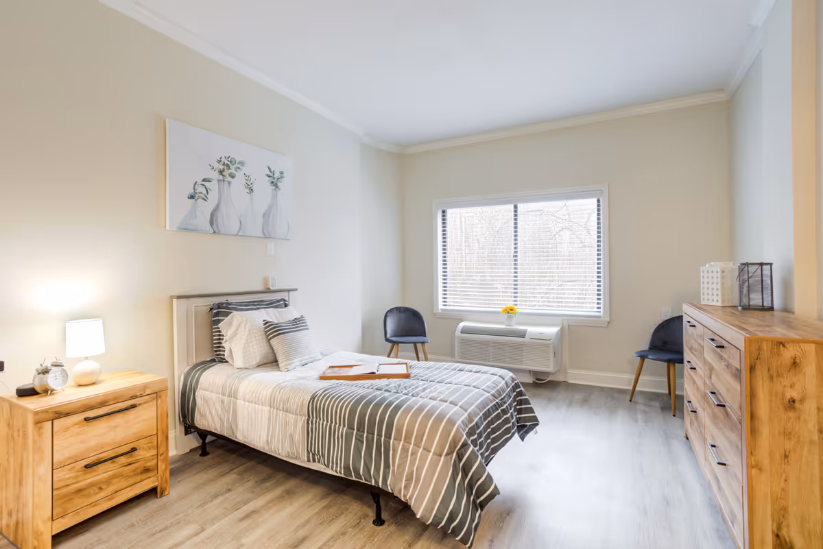 Bright furnished bedroom with a single bed, wooden nightstand and dresser, window with blinds, and two chairs.