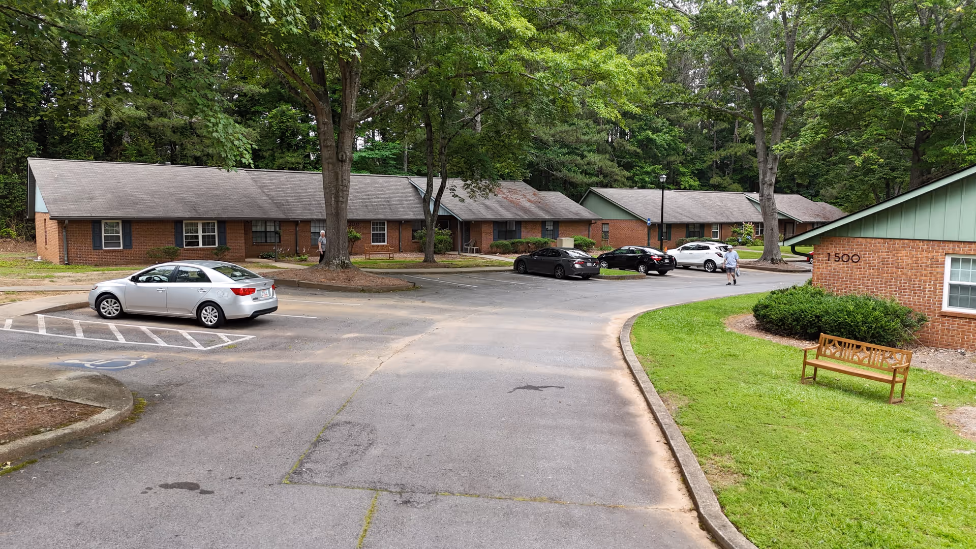 Parking area in front of single-story brick residential buildings surrounded by trees. Several cars are parked, and two people are walking near the buildings. A wooden bench is placed on a grassy area beside one of the buildings.