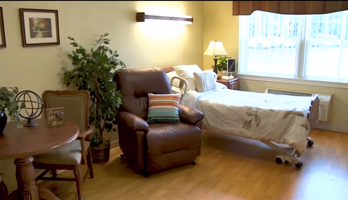 A cozy senior living room featuring a hospital-style bed with white and beige floral bedding near a large window with blinds. Next to the bed is a wooden nightstand with a lamp and a small plant. A brown recliner chair with a striped pillow is positioned beside the bed. There is a round wooden table with two chairs, a decorative plant, and framed artwork on the light yellow walls. The floor is wooden.