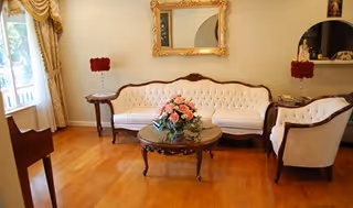 A living room with a vintage-style white tufted sofa and matching armchair, a wooden coffee table with a floral centerpiece, two side tables each holding a red floral arrangement, a large ornate gold-framed mirror on the wall, and a window with heavy drapes allowing natural light to enter.
