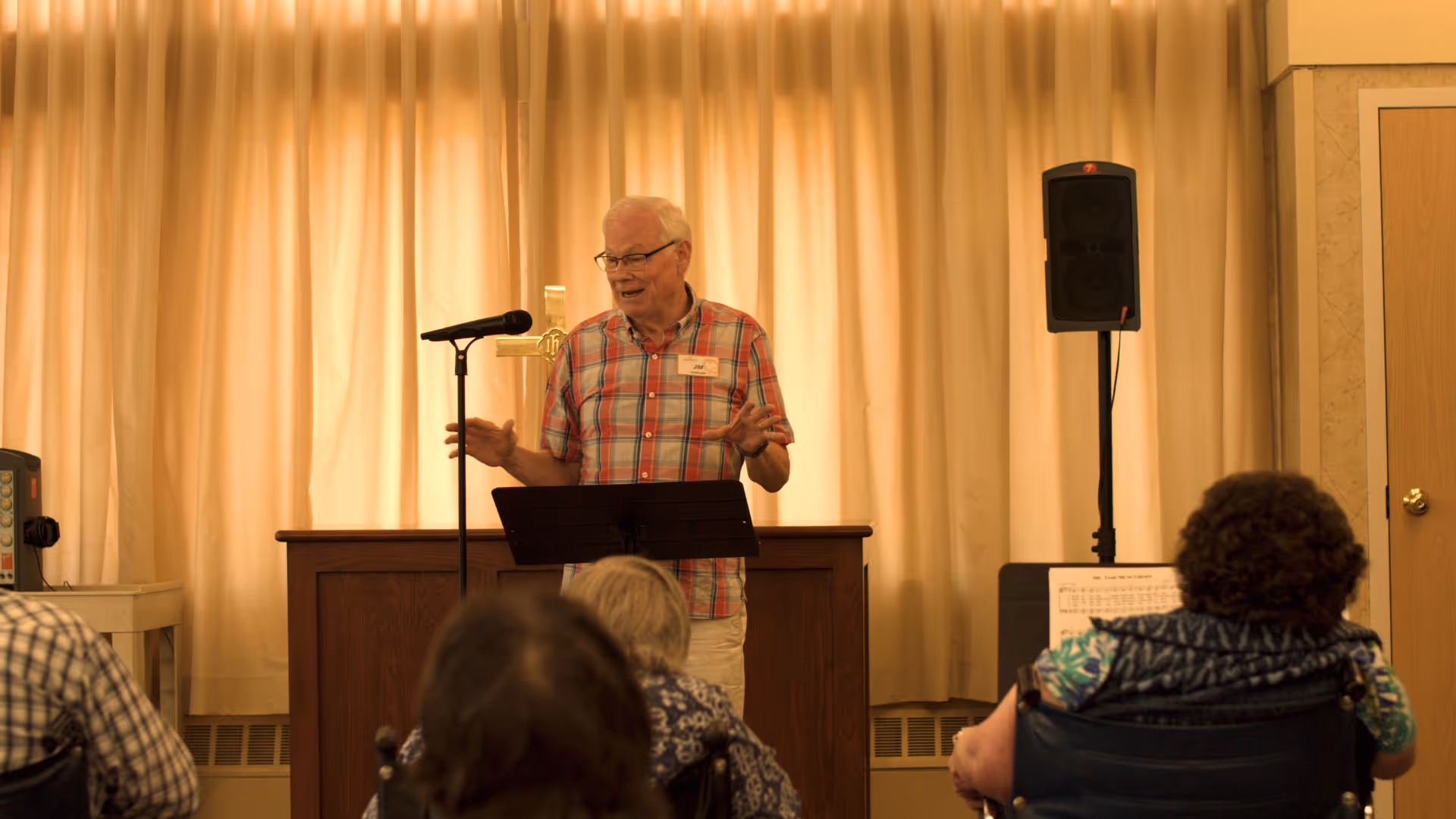 An elderly man wearing a plaid shirt stands behind a podium with a microphone, speaking to an audience seated in front of him in a room with beige curtains and a wooden door.