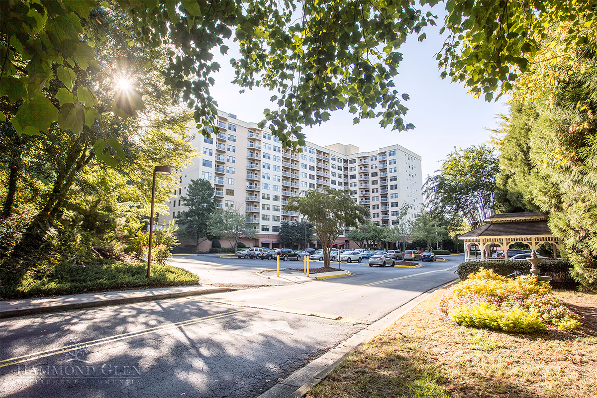 Exterior view of a multi-story retirement community building surrounded by trees and greenery with a parking lot in front. The sun shines through the leaves of a tree on the left side, casting shadows on the road. A gazebo is visible on the right side near landscaped bushes and flowers.