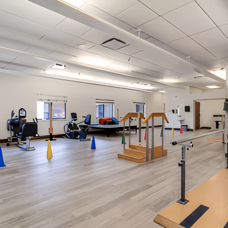 A spacious rehabilitation or physical therapy room with exercise equipment including stationary bikes, parallel bars, a small set of stairs with handrails, and colorful cones placed on the floor. The room has large windows, light-colored wood flooring, and a white ceiling with fluorescent lighting.