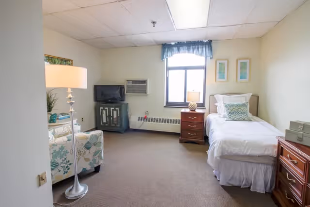 A senior living community room with a single bed dressed in white linens and decorative pillows, a wooden nightstand with a lamp, a window with a blue valance, a small TV on a cabinet, a floral armchair, and a floor lamp.