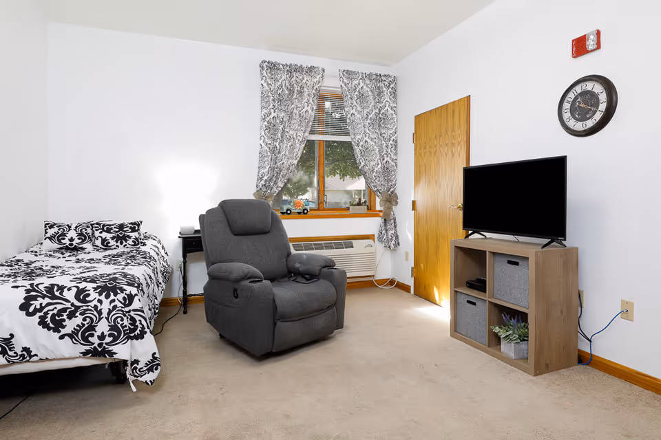 A small, cozy room with a single bed covered in black and white patterned bedding, a gray recliner chair, a wooden cabinet with a flat-screen TV on top, and a window with patterned curtains. There is a wall clock above the TV and a wooden door next to the window.