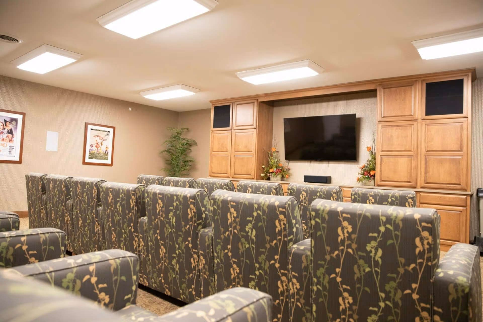 A small theater or media room with several rows of floral patterned armchairs facing a wall-mounted flat screen TV. The room has wooden cabinets on either side of the TV, beige walls with framed posters, and ceiling lights providing bright illumination.