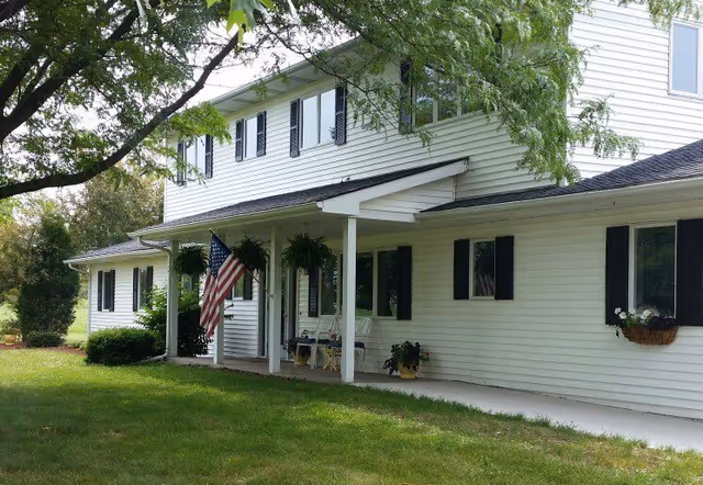 Exterior view of a two-story white building with black shutters, a covered porch with hanging plants, an American flag, and a green lawn with trees in the foreground.