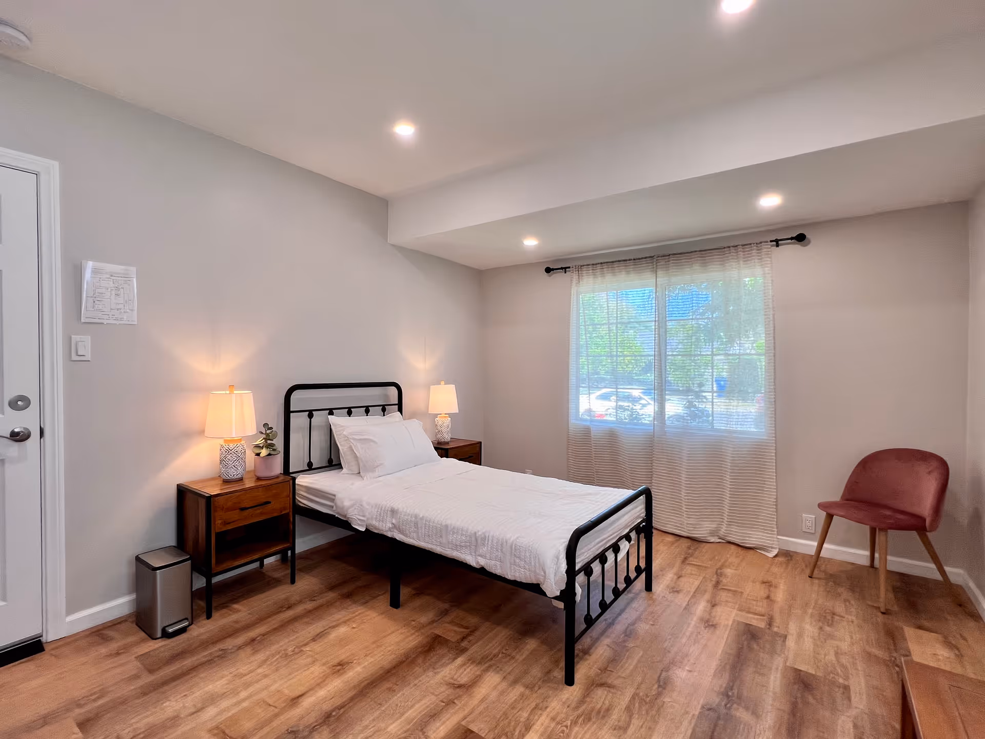 A simple, clean bedroom with a single black metal bed frame, white bedding, two wooden nightstands each with a lamp, a small plant on one nightstand, a pink upholstered chair in the corner, wood flooring, and a large window with sheer curtains letting in natural light.
