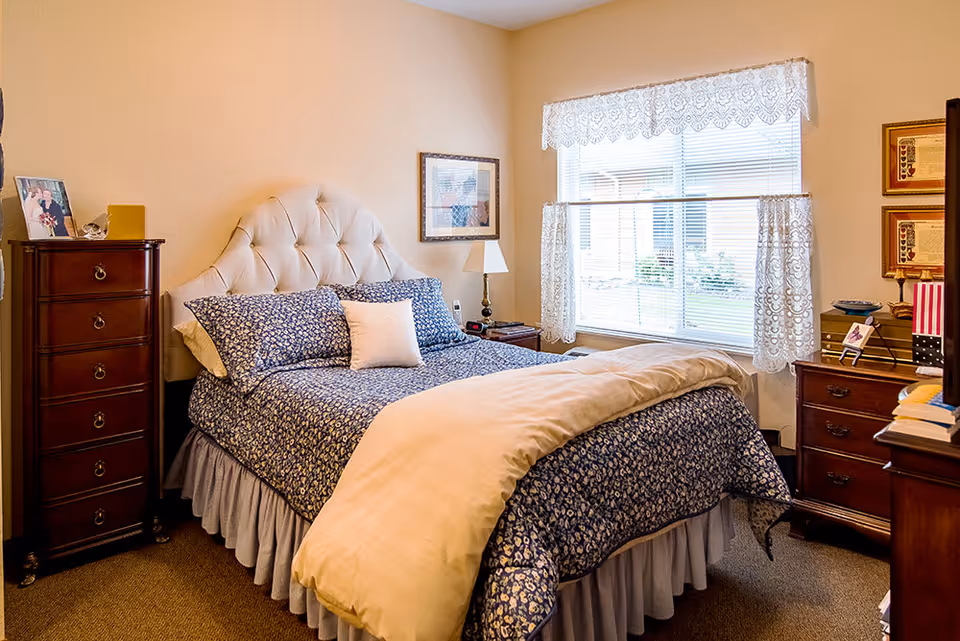 A cozy bedroom with a large bed featuring a tufted headboard, blue floral bedding, and a beige comforter. The room has a tall wooden dresser on the left, a nightstand with a lamp and clock beside the bed, and a window with lace curtains letting in natural light. There are framed pictures on the walls and additional wooden furniture on the right side.
