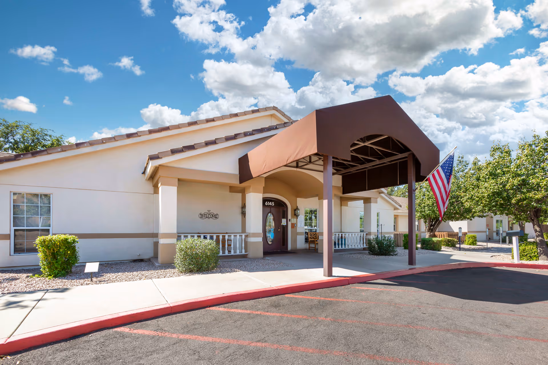 Exterior view of Brookdale East Mesa facility entrance with a covered driveway, American flag, and clear blue sky with clouds.