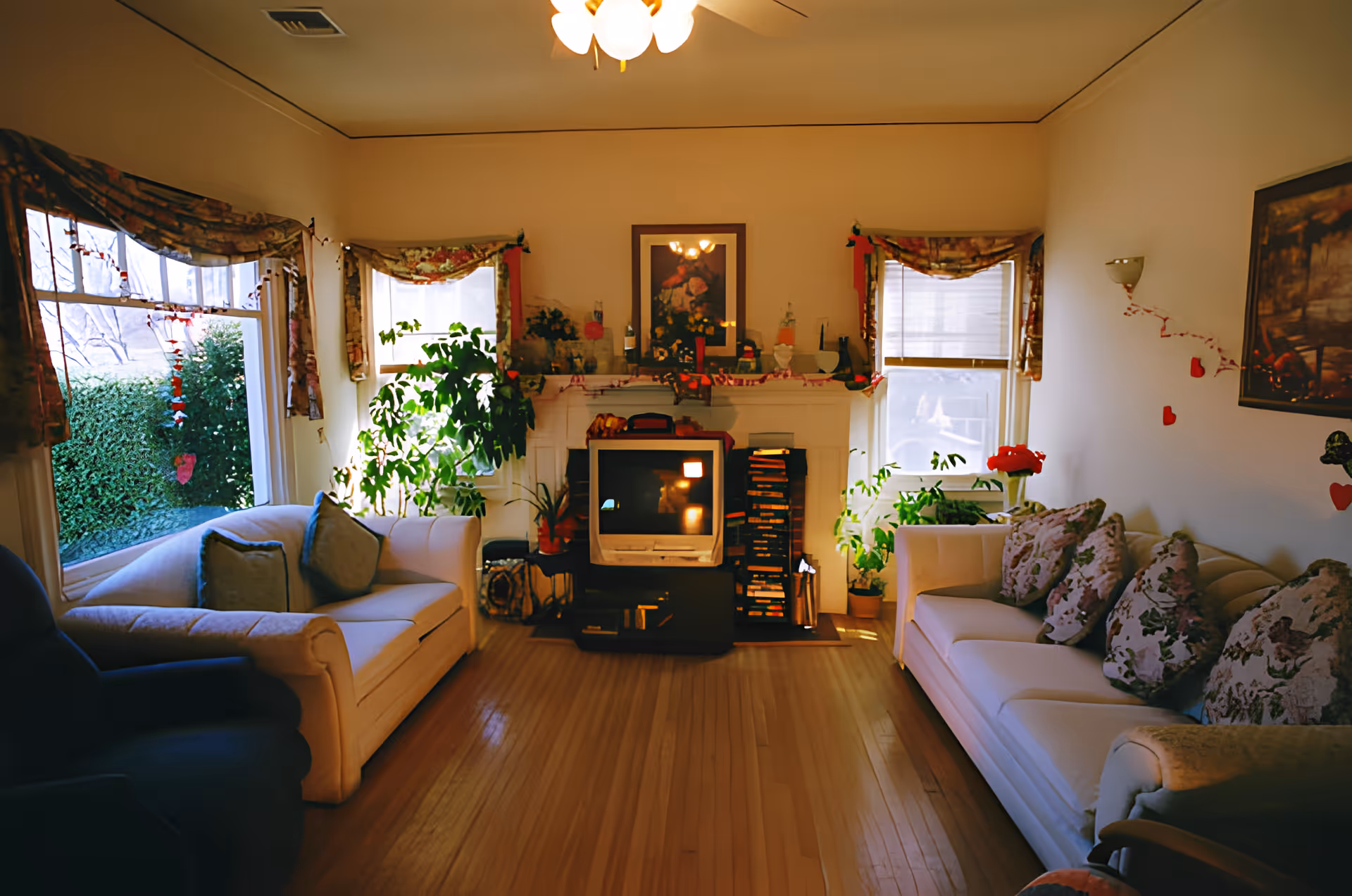 A cozy living room with two white sofas adorned with floral cushions, a dark armchair, and several green plants near two windows with patterned curtains. A vintage television sits on a stand in front of a white fireplace mantel decorated with various items and a framed picture above it. The room has wooden flooring and warm lighting.