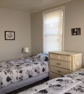 A small bedroom with two twin beds featuring black and white floral bedding. Between the beds is a cream-colored dresser with three drawers. A window with closed blinds is centered on the wall behind the dresser. A small lamp is on a nightstand next to one of the beds. Two framed black and white pictures hang on the walls.