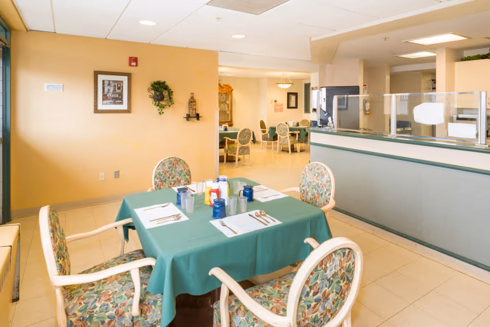 A dining area in a senior living facility with a table covered in a green tablecloth set for four people. The chairs have floral upholstery. In the background, there are more tables and chairs, a serving counter with a glass partition, and warm yellow walls decorated with framed pictures and a wall-mounted plant.