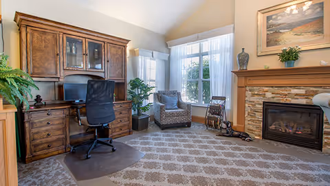 A cozy living room area with a large wooden desk and black office chair on the left, a patterned armchair near a window with white curtains, a fireplace with a stone surround and wooden mantel on the right, and a patterned carpet covering the floor. There are decorative plants and artwork above the fireplace.