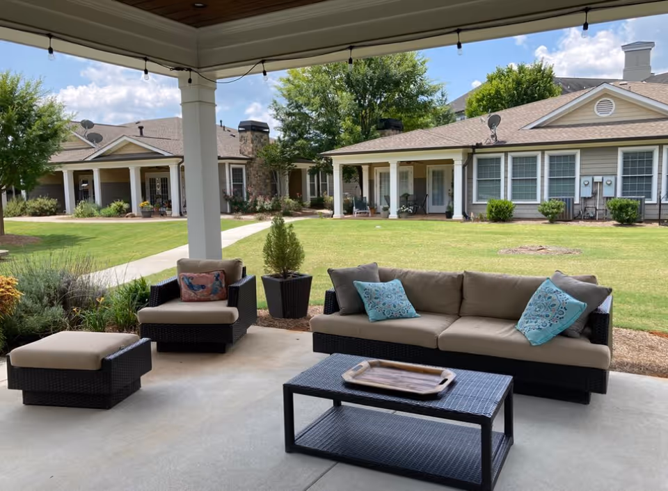 Outdoor covered patio area with cushioned wicker furniture including a sofa, armchair, ottoman, and coffee table with a tray. The patio overlooks a well-maintained lawn and senior living facility buildings with porches and greenery under a partly cloudy sky.