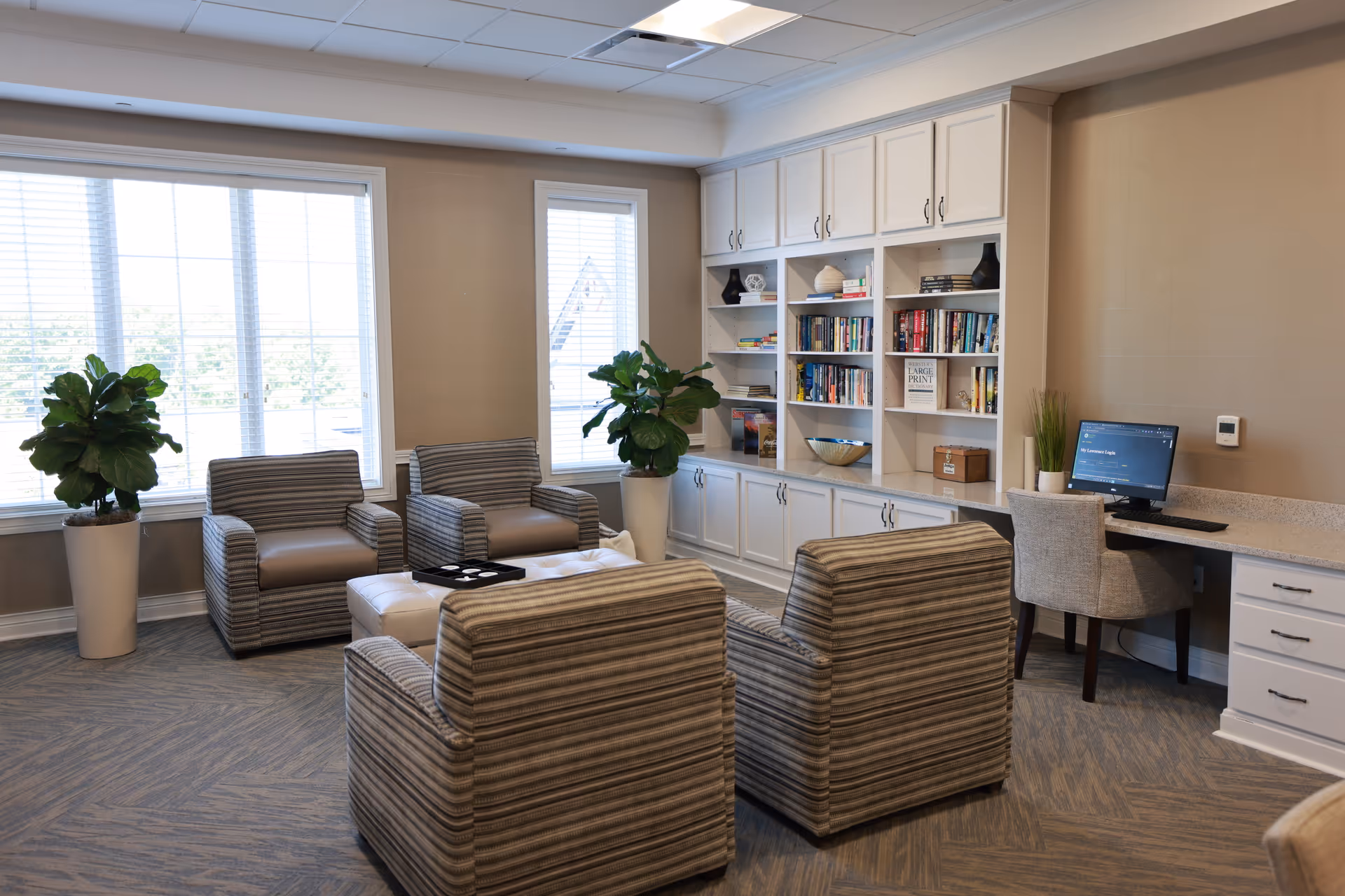 Bright common lounge with striped armchairs around an ottoman, built-in bookshelves, potted plants and a small computer desk.