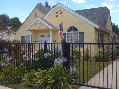Front view of a single-story beige building with a porch, fence, and flowering garden.