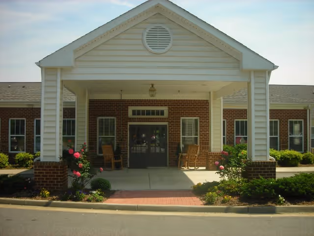 Entrance of a brick building with a covered porch supported by white columns. Two wooden rocking chairs are placed on the porch, surrounded by small bushes and flowering plants. The sky is clear with some light clouds.