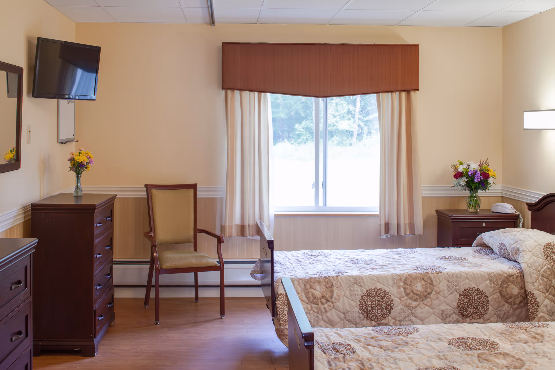A well-lit bedroom in a senior living facility with two twin beds covered in beige and brown patterned quilts. There is a window with beige curtains and a brown valance letting in natural light. The room has wooden furniture including a dresser with a vase of flowers, a nightstand with a telephone and another vase of flowers, and a chair with a cushioned seat. A flat-screen TV is mounted on the wall above the dresser.