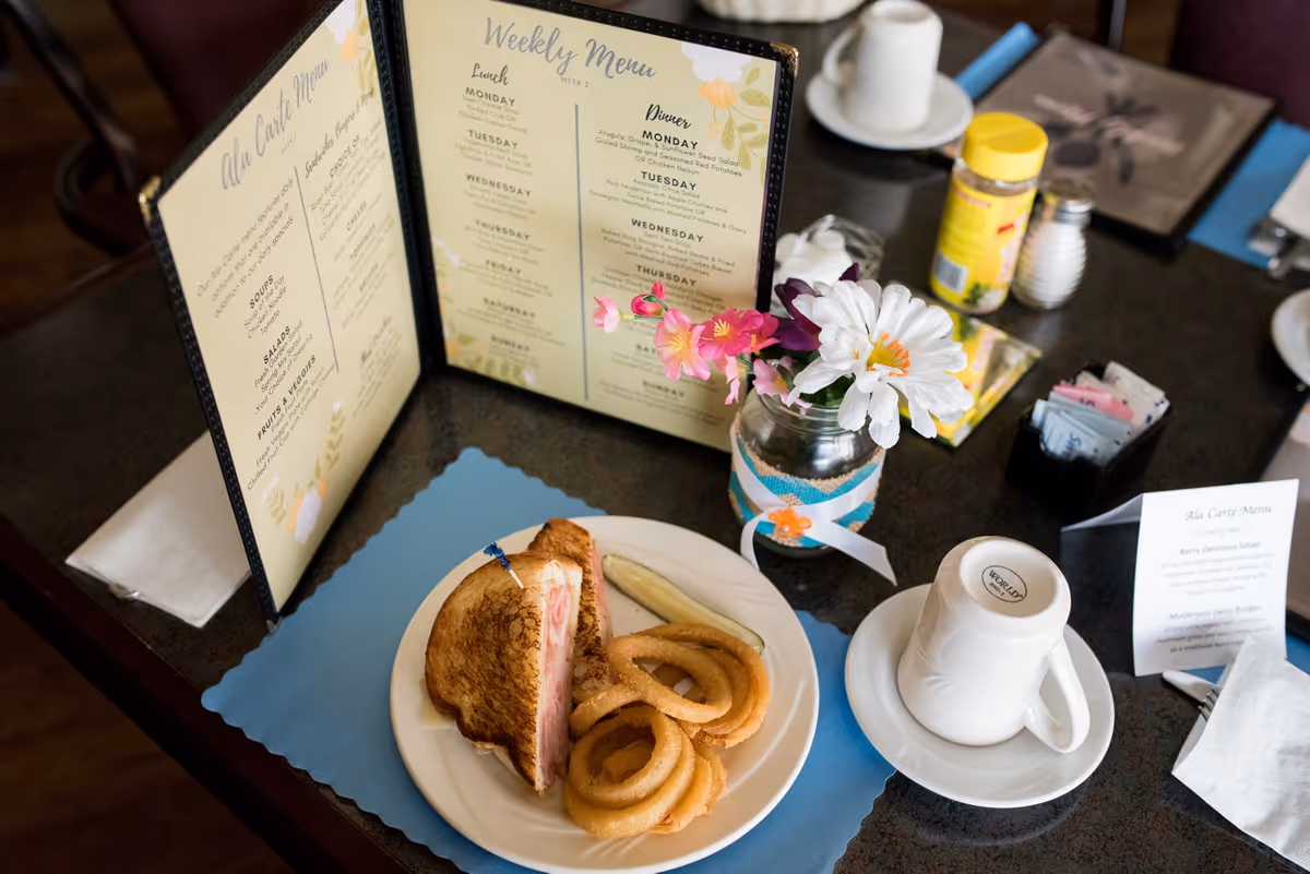 A dining table with a sandwich and onion rings, a weekly menu, and a small vase of flowers.