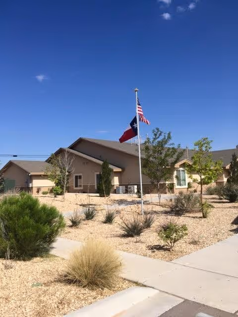 Exterior view of a single-story building with beige walls and a brown roof under a clear blue sky. In front of the building, there is a landscaped area with small trees, bushes, and desert plants, along with a flagpole displaying the American and Texas flags. A concrete sidewalk runs alongside the landscaped area.