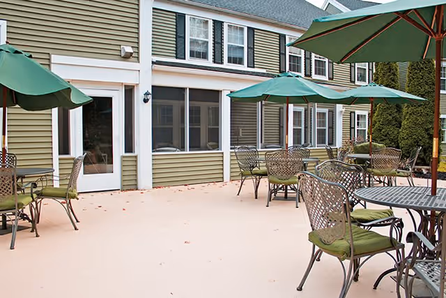 Outdoor patio area with several metal tables and chairs, each table shaded by large green umbrellas, adjacent to a building with beige siding and multiple windows.