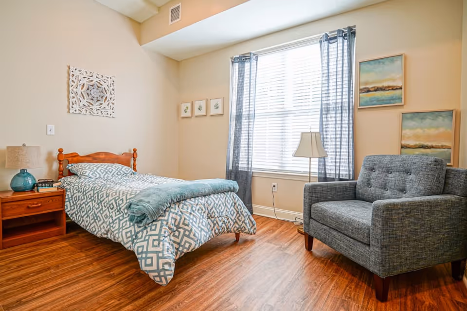A cozy bedroom with a single wooden bed dressed in blue and white patterned bedding and a teal throw blanket. Next to the bed is a wooden nightstand with a blue lamp and a couple of books. A large window with sheer blue curtains lets in natural light. A gray upholstered armchair is positioned near the window, and the walls are decorated with framed artwork. The floor is wooden.