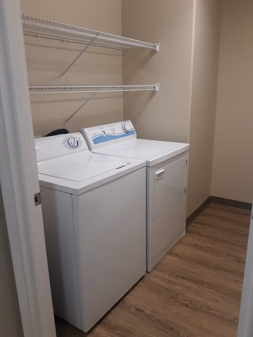 A small laundry room with a white washing machine and dryer side by side, two white wire shelves mounted on the beige wall above them, and wood-look flooring.