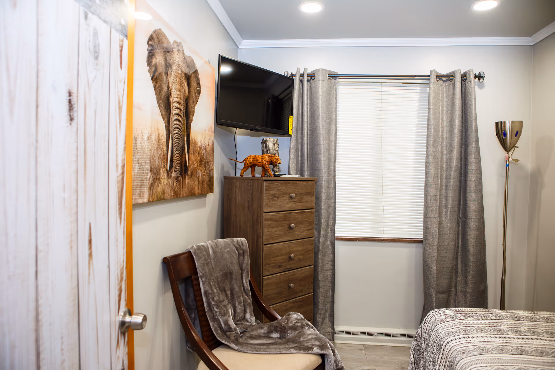 A cozy bedroom featuring a wooden chair with a gray blanket draped over it, a wooden dresser with decorative items including a small leopard figurine, a wall-mounted flat-screen TV, a window with closed blinds and gray curtains, a floor lamp, and a bed partially visible with a patterned bedspread. A large elephant artwork hangs on the wall.