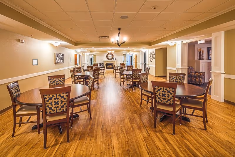 A spacious dining room with multiple round wooden tables and patterned cushioned chairs arranged neatly on a wooden floor. The room features warm lighting, beige walls, and a fireplace with a large clock above it at the far end. There are decorative elements on the walls and a cozy seating area visible through an open doorway on the right.