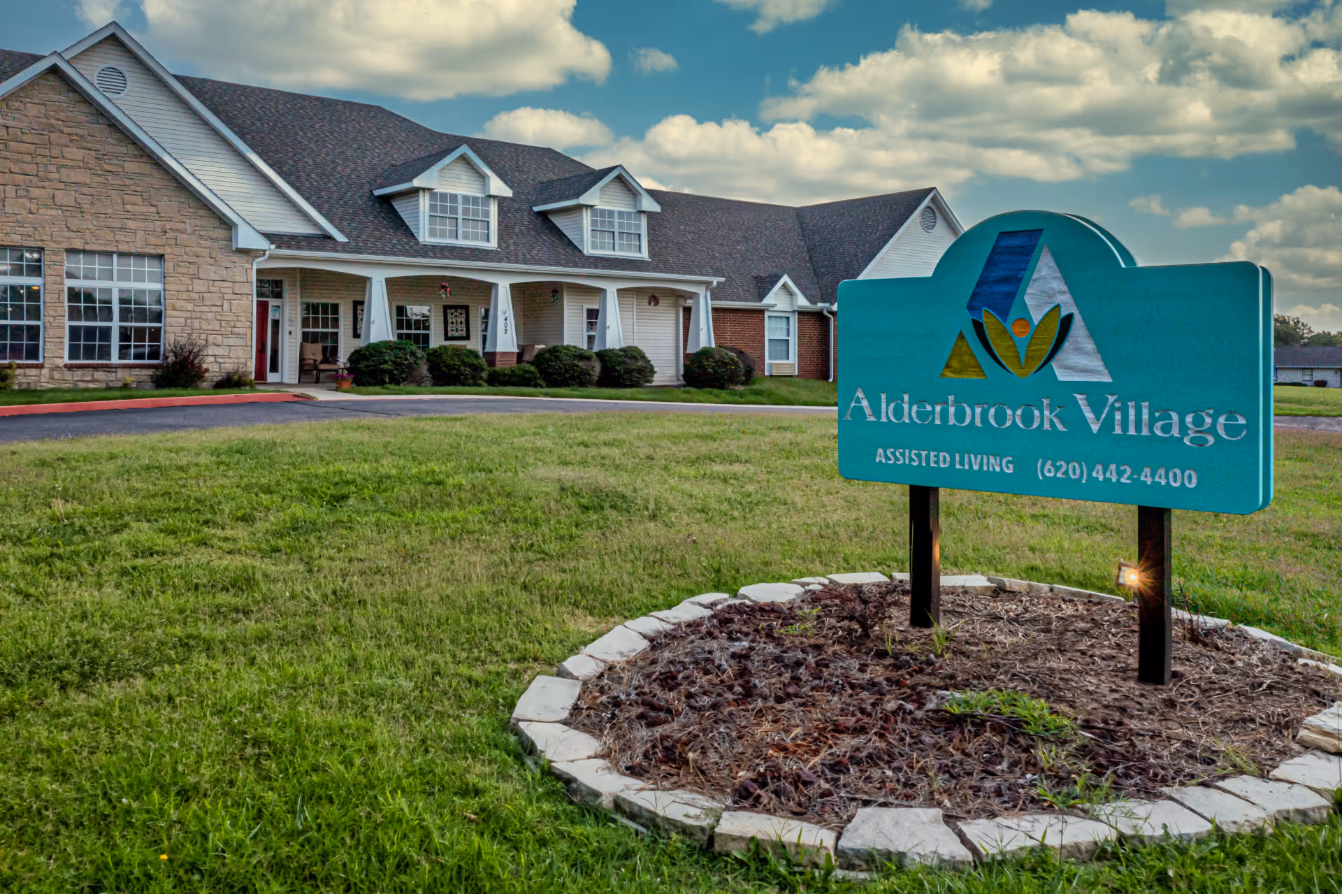 Exterior view of Alderbrook Village assisted living facility showing a large building with a stone and siding facade, multiple windows, and a covered porch. In the foreground, there is a green sign with the facility's name, logo, and contact number, surrounded by a small landscaped area with mulch and stones. The sky is partly cloudy.