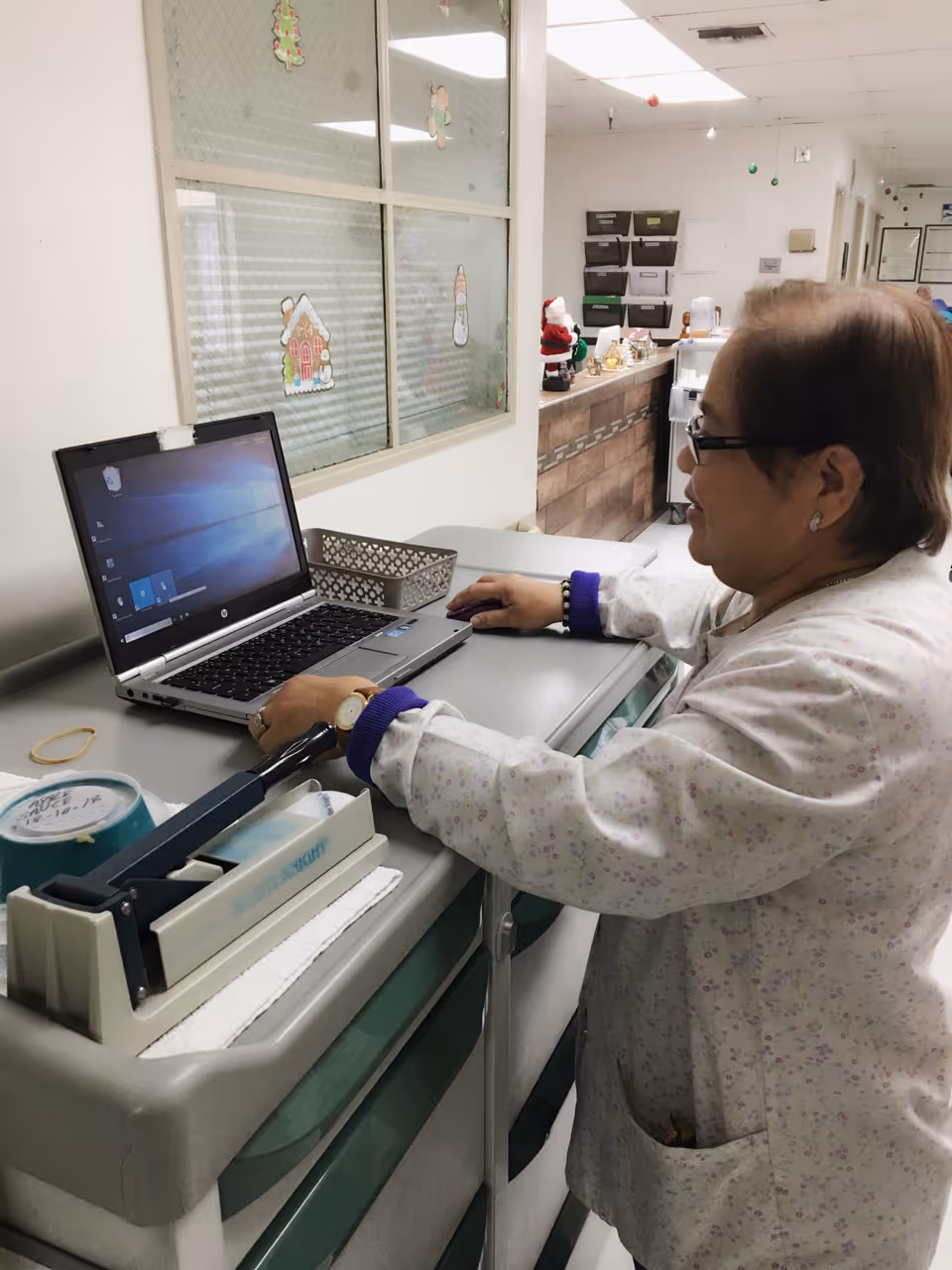 A woman wearing glasses and a light-colored patterned jacket is standing at a medical cart using a laptop computer in a hallway decorated with Christmas-themed window clings and small holiday decorations on a counter in the background.