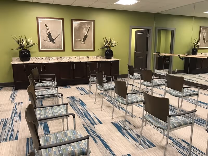 Room with green walls and a patterned carpet featuring blue and beige stripes. There are two rows of chairs with metal frames and striped cushions facing a mirrored wall with a ballet barre. A long cabinet with a granite countertop is against the wall, decorated with two black vases holding plants and two framed pictures of divers in mid-air above it.
