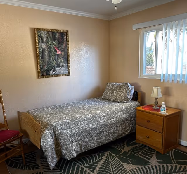 A small bedroom with a single bed covered in a patterned gray and white bedspread. Next to the bed is a wooden nightstand with two drawers, a small lamp, a bottle of hand sanitizer, and a red cloth. A wooden chair with a red cushion is positioned near the bed. A window with vertical blinds allows natural light into the room. A framed picture hangs on the beige wall above the bed. The floor has a carpet with a large leaf pattern.