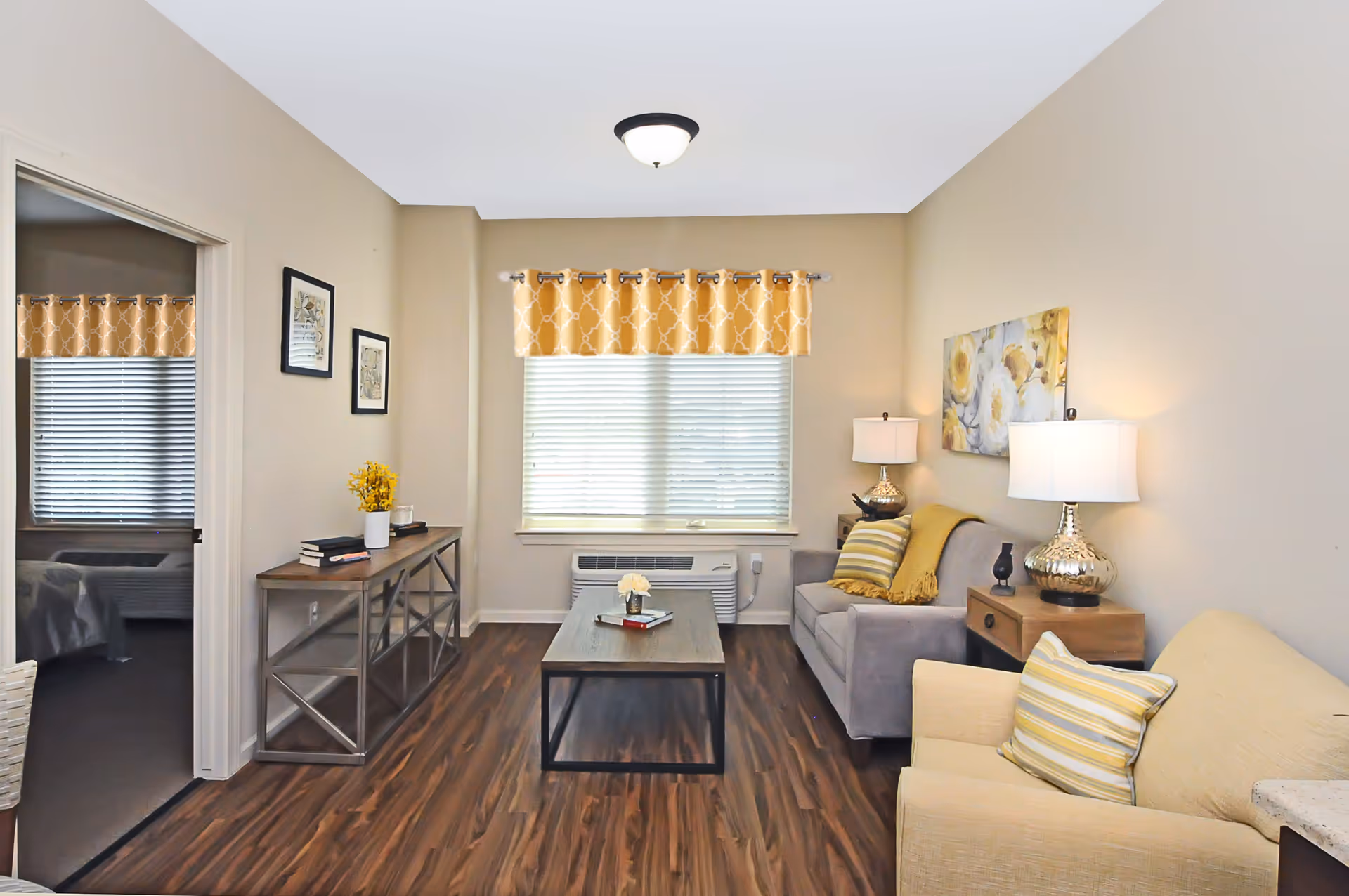 A cozy living room with wood flooring, a gray sofa with yellow and striped pillows, a beige armchair with a striped pillow, a wooden coffee table with a small flower vase and books, two table lamps on wooden side tables, a window with yellow patterned valance, and a console table with books and a flower vase. A doorway leads to a bedroom with a bed and window with matching yellow valance.