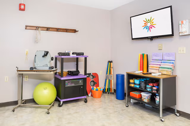 A therapy or activity room with exercise and therapy equipment including a large green exercise ball, a purple cart with electronic devices, colorful foam rollers, a blue foam cylinder, a bucket with balls, and a small cabinet with various supplies. A flat screen TV is mounted on the wall displaying a colorful logo.