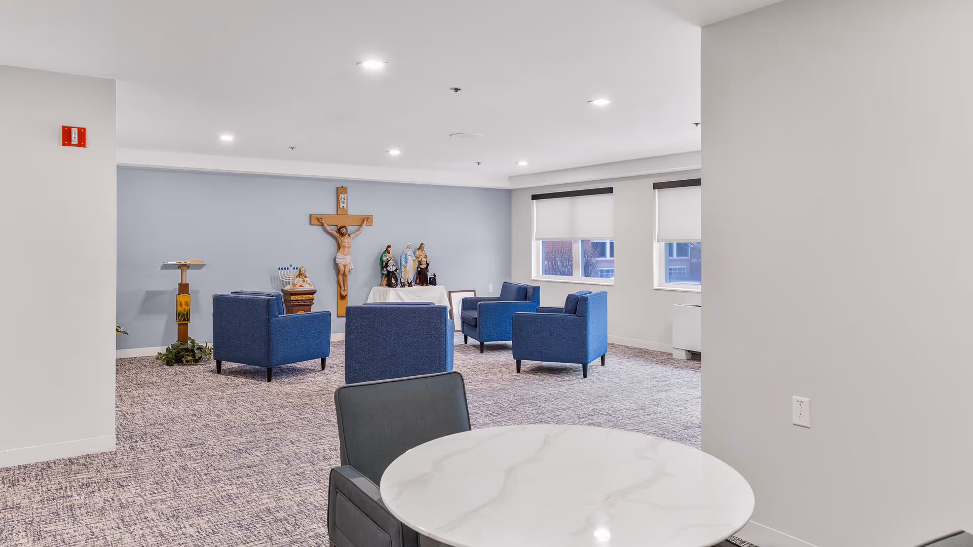 A quiet room with blue armchairs arranged in a semi-circle facing a religious altar featuring a crucifix and several statues. The room has light gray walls, carpeted floor, and two windows with white blinds partially drawn. A round white marble table with a dark chair is in the foreground.