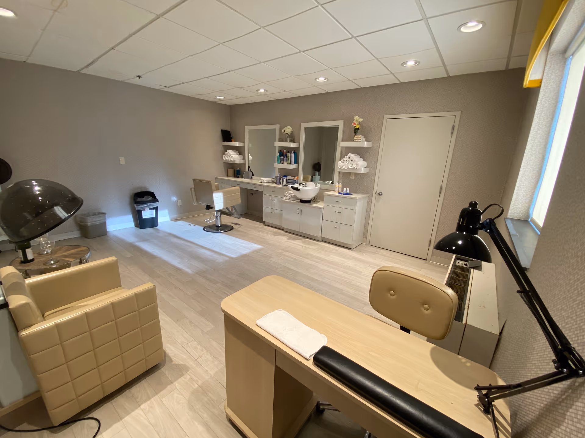 A well-lit salon room with beige walls and light wood flooring. The room contains a salon chair in front of a counter with mirrors, shelves stocked with towels and hair products, a hair dryer hood, a beige armchair, and a manicure table with a lamp and a towel. There is a closed door and a window with blinds on the right side.