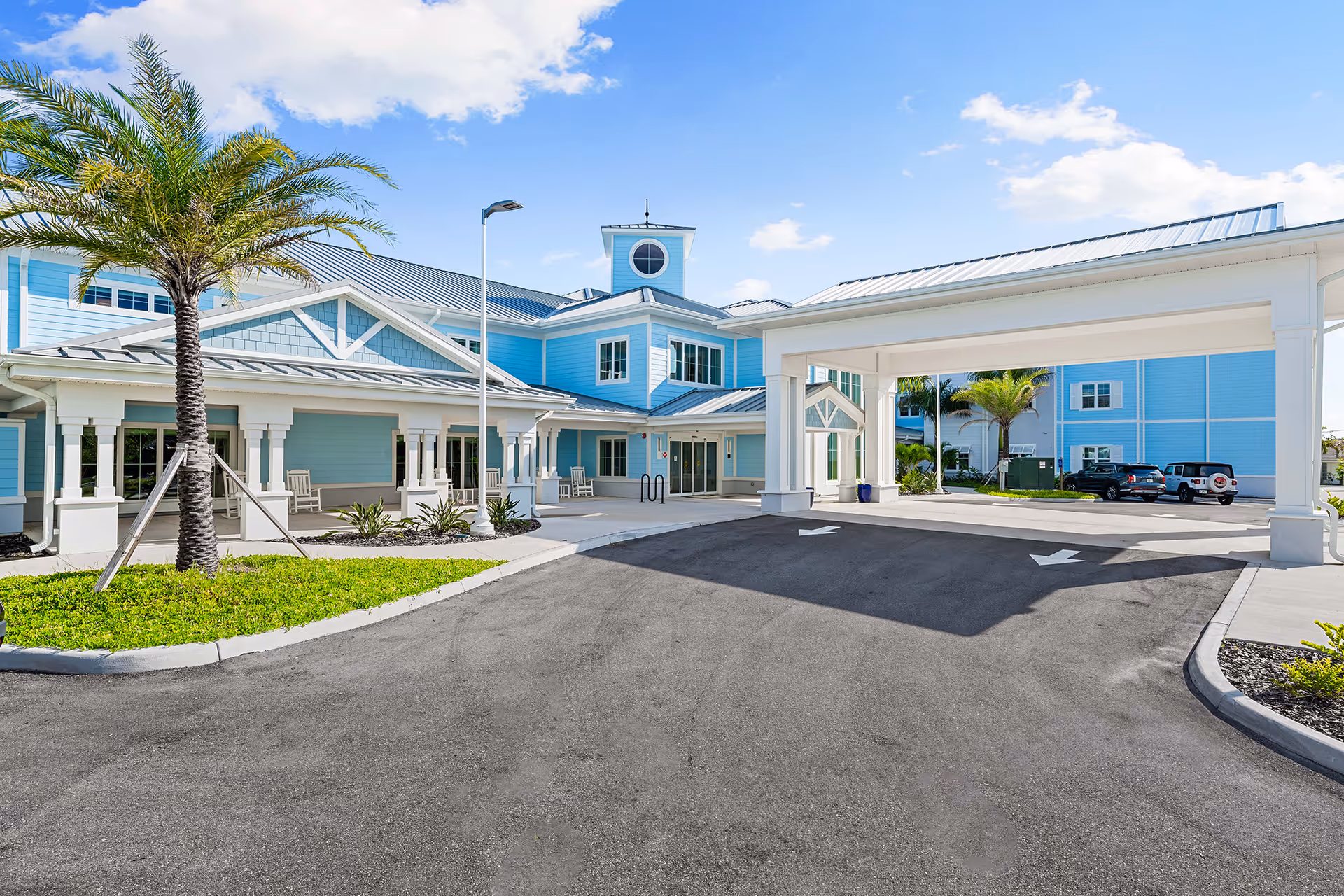 Exterior view of a senior living facility with light blue buildings, a covered entrance driveway, palm trees, and a clear blue sky.