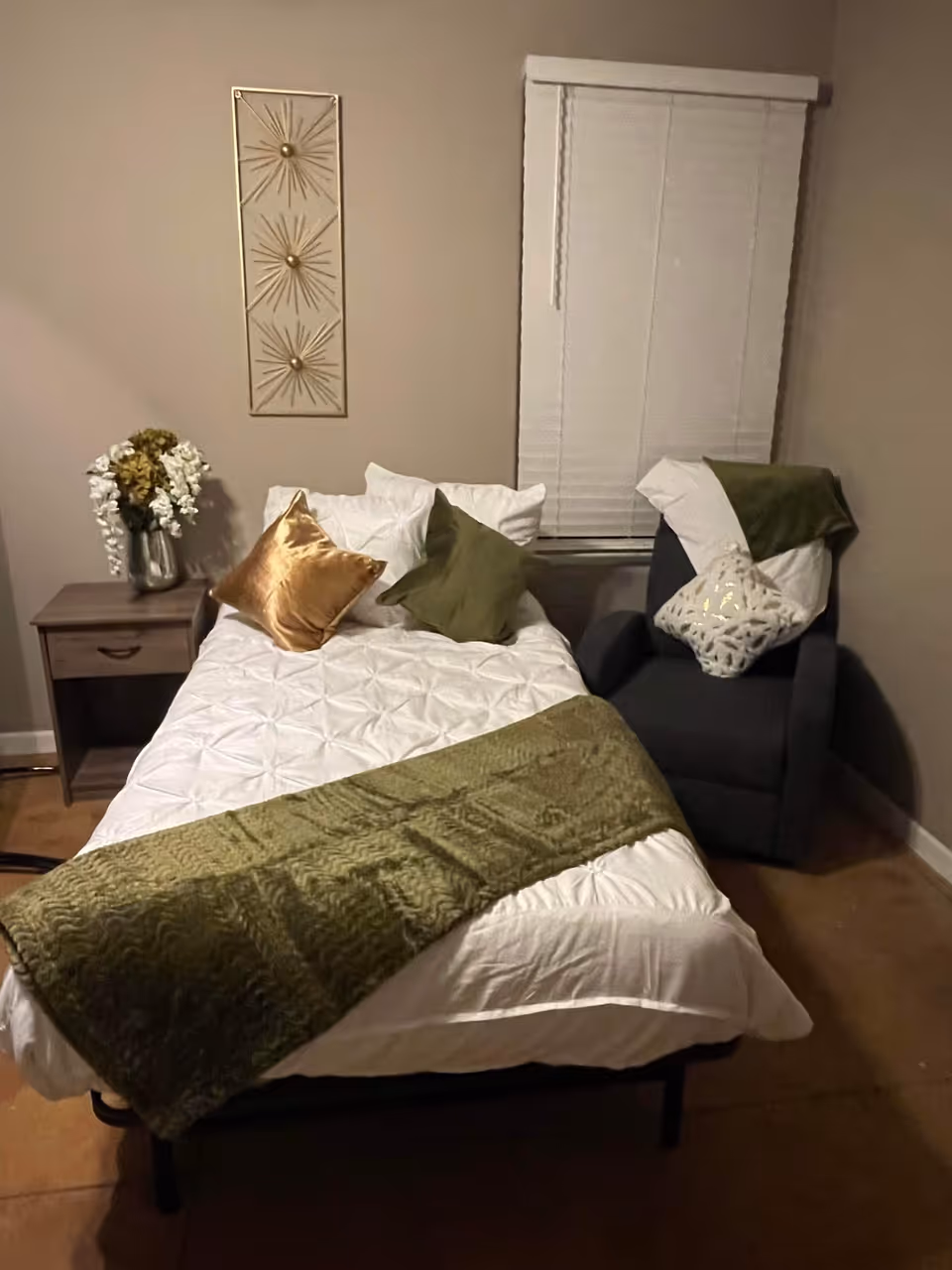 A small bedroom with a single bed made with white bedding, green throw and decorative pillows, a nightstand with flowers, and an armchair beside a window with blinds.