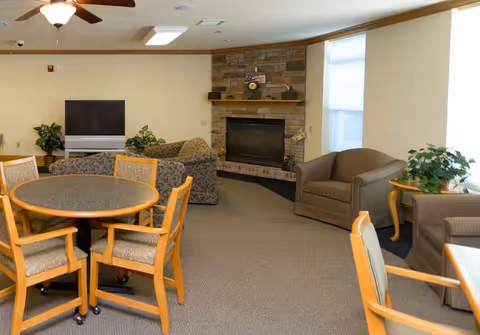 A senior living facility common area featuring a round table with four wooden chairs in the foreground, a stone fireplace with a mantel decorated with small items, two armchairs, a patterned sofa, a flat-screen TV on a stand, and potted plants near the windows.