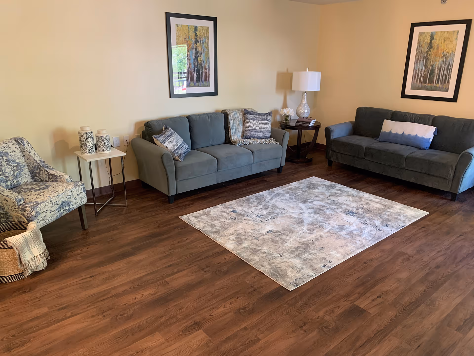 A living room with two gray sofas, an upholstered armchair, side tables, a lamp, framed artwork, and a rectangular area rug on wood floors.